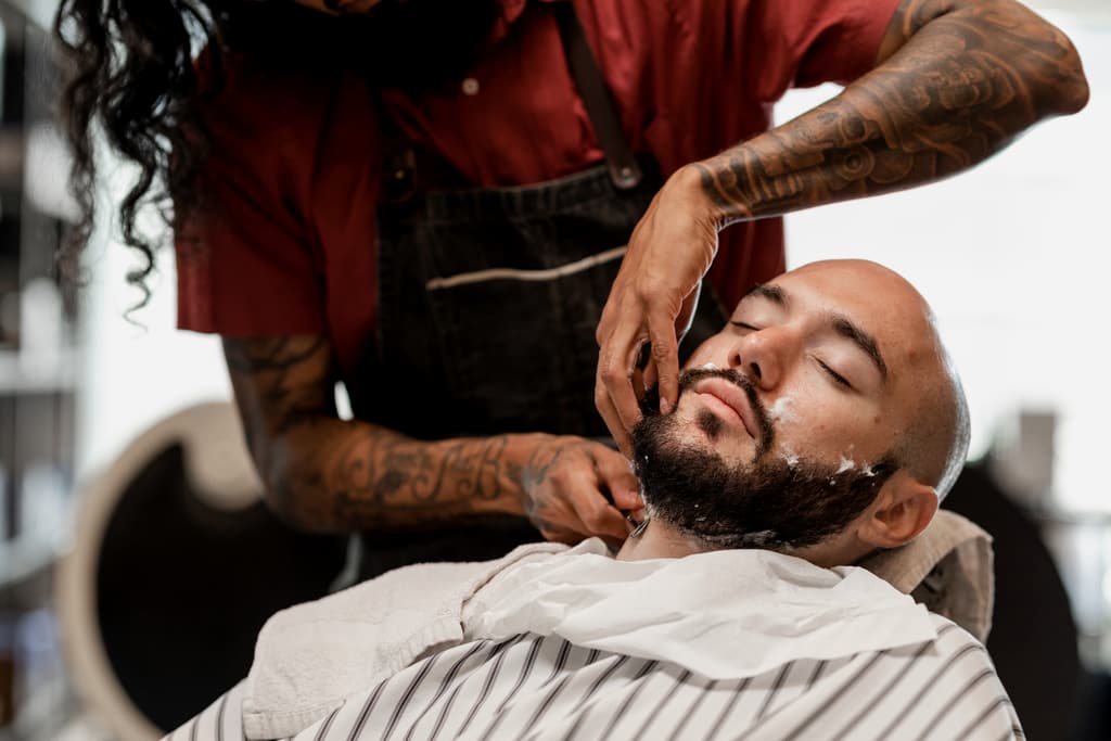 Un barbero de brazos tatuados prepara la barba de un cliente para el afeitado.