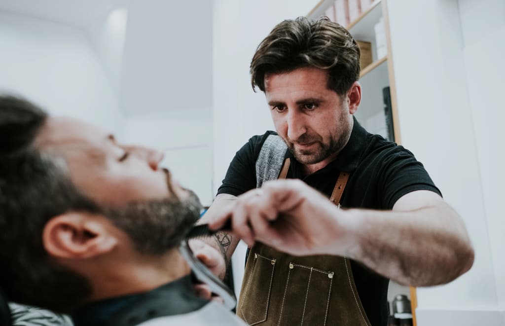 Un barbero perfila a tijera la barba de un cliente recostado.