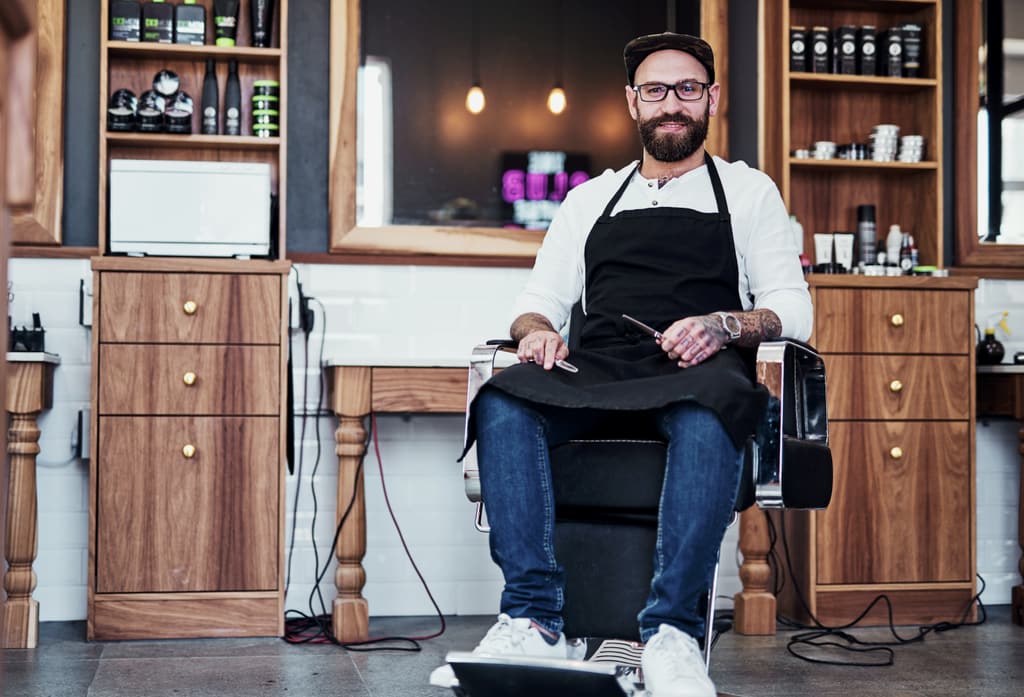 Barbero con delantal sentado en el sillón de su barbería, entre muebles de madera.